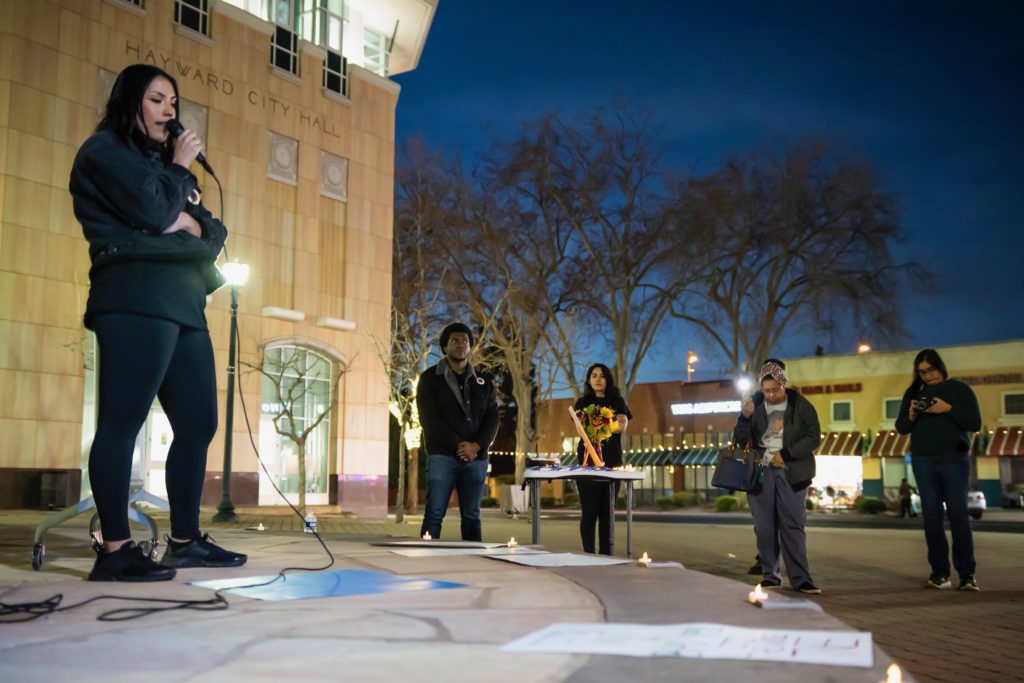 One organizer on stage in front of City Hall speaking while other organizers and audience members listen.