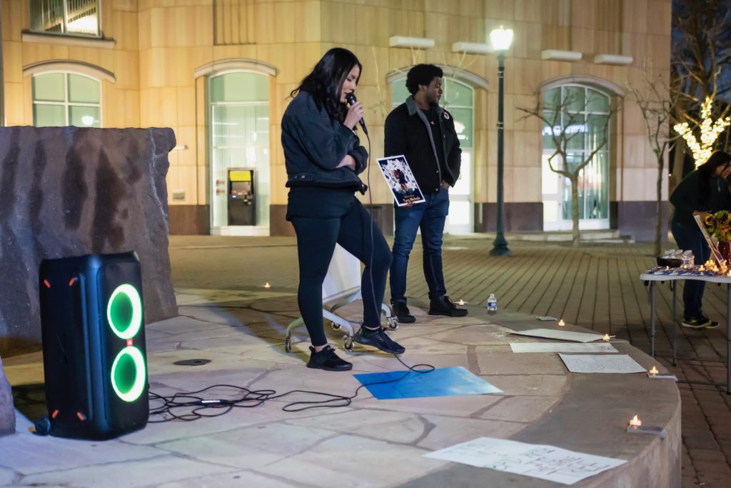 Two people standing on stage, one is speaking and the other is holding a sign dedicated to Tyre Nichols.