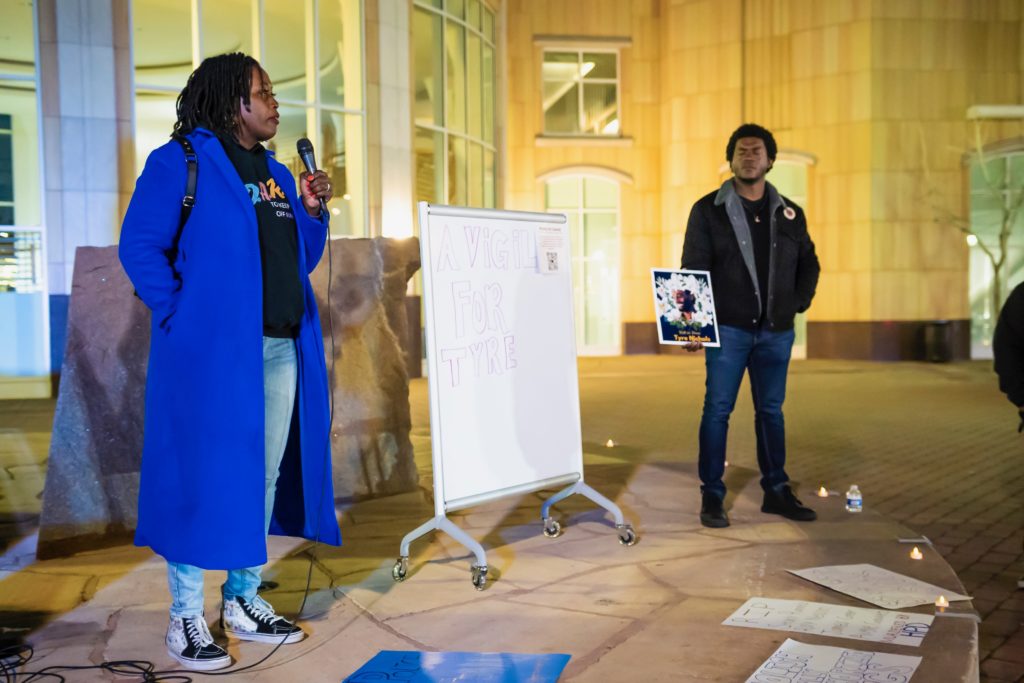 Two people on stage, one speaking and another is holding a sign dedicated to Tyre Nichols.