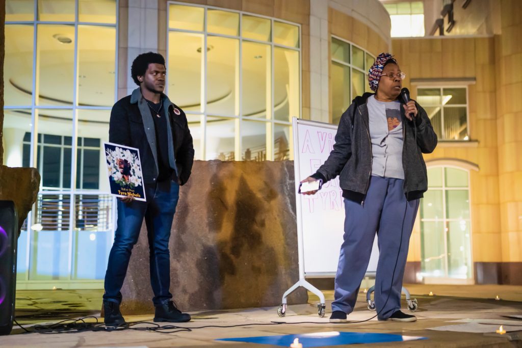 Two people on stage in front of City Hall. One is giving a speech and the other is holding a sign dedicated to Tyre Nichols.